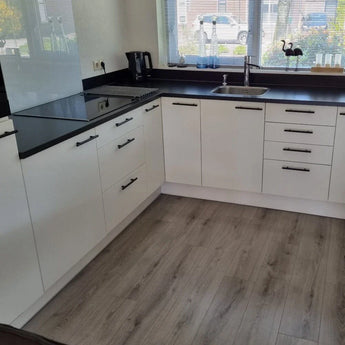 Moderne keuken met witte kasten met yessential.nl Remo Black handgreep, zwart aanrechtblad, elektrische kookplaat, ingebouwde oven, koffiezetapparaat en een gootsteen onder een raam. Lichthouten vloeren en kleine voorwerpen sieren de vensterbank.