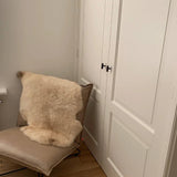 Een moderne beige stoel met een crèmekleurig bont kleedje staat naast witte kastdeuren met panelen en Vasco Black Knobs van yessential.nl in een matte afwerking, tegen lichte muren en een houten vloer.
