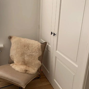 Een moderne beige stoel met een crèmekleurig bont kleedje staat naast witte kastdeuren met panelen en Vasco Black Knobs van yessential.nl in een matte afwerking, tegen lichte muren en een houten vloer.