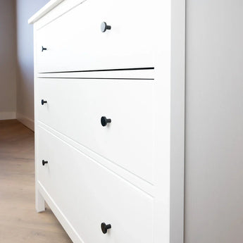 Een close-up van een wit dressoir met drie lades, elk versierd met twee Marizo Black knoppen van yessential.nl. Het dressoir staat op een lichthouten vloer naast een lichtgrijze muur, wat het minimalistische ontwerp benadrukt.