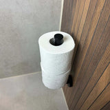 De Roberto Black reserverolhouder van yessential.nl is verticaal gemonteerd op een badkamermuur met beige tegels en houten lambrisering en houdt twee rollen toiletpapier vast.