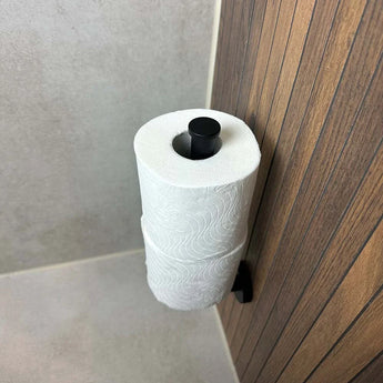 De Roberto Black reserverolhouder van yessential.nl is verticaal gemonteerd op een badkamermuur met beige tegels en houten lambrisering en houdt twee rollen toiletpapier vast.