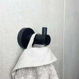 De Donato Black WC Set van yessential.nl heeft een matzwarte wandhaak die een handdoek met witte textuur vasthoudt tegen lichtgrijze tegels, en voegt moderne stijl toe aan je badkameraccessoires.