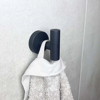 Een lichtgrijze handdoek met structuur hangt met een lus aan een zwart metalen haakje van de yessential.nl Ferro Black WC-set, gemonteerd tegen een effen, licht betegelde muur.