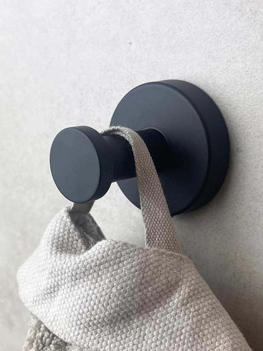 Een ronde, matzwarte wandhaak uit de Xavo Black WC set van yessential.nl houdt een lichtgrijze handdoek of tas met textuur tegen een lichtgekleurde muur.