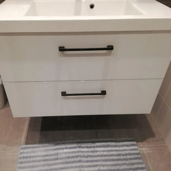 Een modern Ramiro Black Deluxe badmeubel van yessential.nl, met chique zwarte handgrepen, is gemonteerd op een betegelde muur boven een grijs en wit gestreept tapijt, met de rand van een wastafel zichtbaar bovenop.
