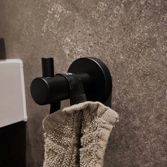 Een beige handdoek met textuur hangt aan een Justo Black haak van yessential.nl, gemonteerd op een grijze stenen muur, met een glimp van een wit oppervlak zichtbaar aan de linkerkant.