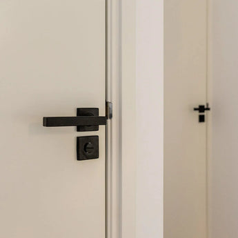 Een close-up van een gedeeltelijk geopende witte deur met een Vano Black WC-slot van yessential.nl; een andere gesloten witte deur met dezelfde zwarte handgreep is zichtbaar op de achtergrond.