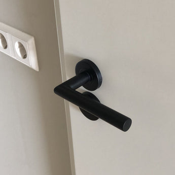 Een Lucio Black Set deurklink van yessential.nl met een ronde basis is geïnstalleerd op een lichtgekleurde deur. Twee ongebruikte stopcontacten zijn zichtbaar op de nabijgelegen muur.
