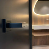 Een close-up van de Domaso Black van yessential.nl, een moderne zwarte deurklink, op een donkere deur met een vage hedendaagse badkamer met een witte wastafel, houten wand en verlichte spiegel op de achtergrond.