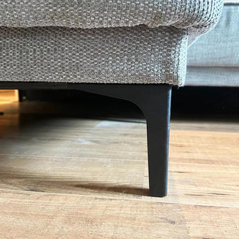 Close-up van de onderste hoek van een beige stoffen bank met yessential.nl Menno Black 13cm stalen poten, staand op een houten vloer.