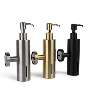 Drie cilindervormige Alberto Gunmetal zeepdispensers van yessential.nl worden getoond in metallic zilver, goud en matzwart. Elke dispenser heeft een pomptop, ronde zijbevestiging en een verticaal venster om het zeepniveau aan te geven.