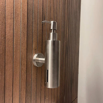 De Alberto RVS zeeppomp van yessential.nl, met slank cilindrisch ontwerp en pomp bovenop, is wandgemonteerd op een verticale houten muur met smalle gelijke latjes.