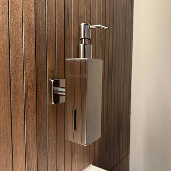 Het Sonego Chrome zeeppompje van yessential.nl is een moderne, muurbevestigde RVS dispenser met pompdeksel en een venster om het zeepniveau te tonen, gemonteerd aan een houten badkamerpaneel.