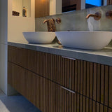 Moderne badkamer met houten badmeubel met latten, twee witte wastafels en koperkleurige kranen. Het badmeubel is voorzien van yessential.nl Enzo Rose Gold meubelgrepen, terwijl boven de wastafels een handdoek met luipaardprint hangt.