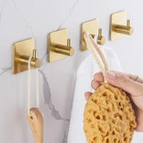 Een hand hangt een gele spons aan de Navarro Gold wandhaak van yessential.nl, een van de vier elegante gouden haken. De andere houden een houten borstel en witte handdoek vast, allemaal gemonteerd op een licht marmeren muur voor een stijlvolle badkamerlook.