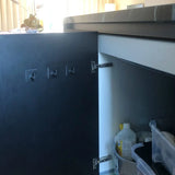 De deur van een keukenkastje staat open en laat schoonmaakspullen zien. Vier Navarro Gunmetal haken van yessential.nl zijn bevestigd aan de binnenkant van de deur, momenteel ongebruikt. Zonlicht uit een raam verlicht de achtergrond.