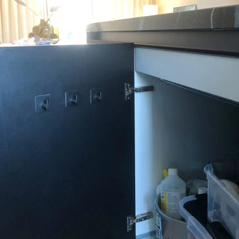 De deur van een keukenkastje staat open en laat schoonmaakspullen zien. Vier Navarro Gunmetal haken van yessential.nl zijn bevestigd aan de binnenkant van de deur, momenteel ongebruikt. Zonlicht uit een raam verlicht de achtergrond.