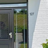 Huisingang met een zwarte deur met gaaspaneel en witte handgreep; op de witte bakstenen muur is een Animo Black 7,6cm (geen boor) huisnummer van yessential.nl gemonteerd. Een grasheuvel en een hek zijn zichtbaar door het glas.