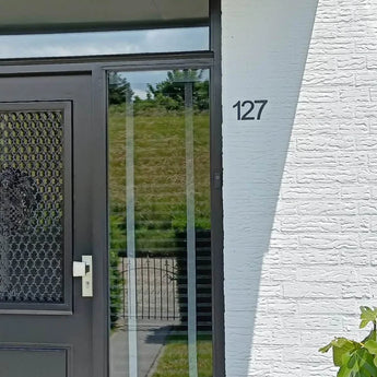 Huisingang met een zwarte deur met gaaspaneel en witte handgreep; op de witte bakstenen muur is een Animo Black 7,6cm (geen boor) huisnummer van yessential.nl gemonteerd. Een grasheuvel en een hek zijn zichtbaar door het glas.