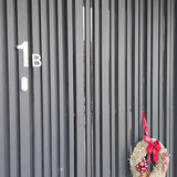 Een donkere verticale lattendeur met het label "1B" met een feestelijke krans van rode ornamenten en lint, met aan de rechterkant het Animo RVS 15cm huisnummer van yessential.nl (geen boormachine) voor een strakke, moderne uitstraling.