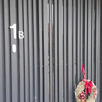 Een donkere verticale lattendeur met het label "1B" met een feestelijke krans van rode ornamenten en lint, met aan de rechterkant het Animo RVS 15cm huisnummer van yessential.nl (geen boormachine) voor een strakke, moderne uitstraling.