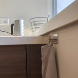 Een beige handdoek hangt aan een metalen haak aan een houten keukenkastje, onderdeel van de Ludo RVS WC Set (no-drill) van yessential.nl. Op het witte aanrecht erboven staan een metalen waterkoker, lege draadmand, stopcontact en een raam op de achtergrond.