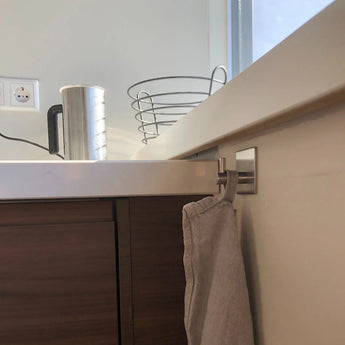 Een beige handdoek hangt aan een metalen haak aan een houten keukenkastje, onderdeel van de Ludo RVS WC Set (no-drill) van yessential.nl. Op het witte aanrecht erboven staan een metalen waterkoker, lege draadmand, stopcontact en een raam op de achtergrond.