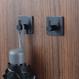 Twee Navarro Black haken van yessential.nl zijn gemonteerd op hout. Aan de ene haak zit de lus van een gesloten zwarte paraplu met een grijs handvat; de andere haak is leeg.