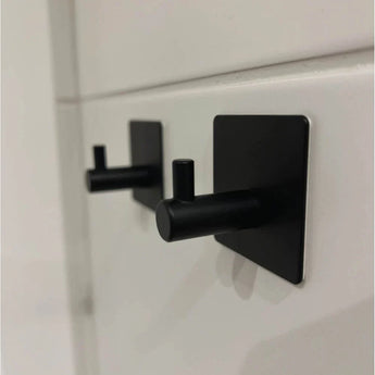 Vier 4x Navarro Black handdoekhaakjes van yessential.nl, met een modern, minimalistisch metalen ontwerp, zijn gemonteerd op een wit betegelde muur.