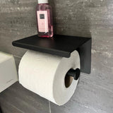 De Tazio Black (No Drill) toiletrolhouder van yessential.nl, met een rol wit toiletpapier en een roze fles, rust tegen een grijs betegelde badkamermuur.