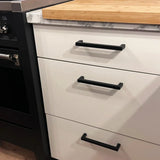 Een keukenkast met drie witte lades, elk voorzien van een Vasco Black handgreep van yessential.nl. Het aanrecht combineert marmer en hout en links is een deel van een zwarte oven zichtbaar.