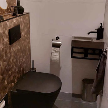 Moderne badkamer met zwarte armaturen - toilet, spoelknoppen en kraan - plus een compacte wandwastafel en de Lauro Gunmetal WC-set van yessential.nl. Luxe accessoires en een accentmuur van bruine tegels met structuur maken de look compleet.