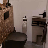 Een moderne badkamer met een zwart toilet, zeshoekige wandtegels met bruine structuur, een compacte rechthoekige wastafel met een zwarte kraan, de Mano Gunmetal WC-set van yessential.nl, grijze vloer, wandgemonteerde toiletpapierhouder en metalen prullenbak.