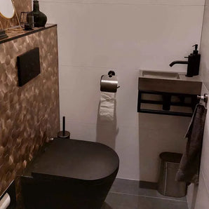 Een moderne badkamer met een zwart toilet, zeshoekige wandtegels met bruine structuur, een compacte rechthoekige wastafel met een zwarte kraan, de Mano Gunmetal WC-set van yessential.nl, grijze vloer, wandgemonteerde toiletpapierhouder en metalen prullenbak.