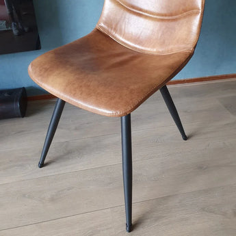 Een moderne stoel met een lichtbruine kunstleren zitting en zwarte metalen poten, voorzien van yessential.nl Paco Set Ø 17-21mm anti-kras stoelpoot beschermers, staat op een houten vloer tegen een blauwe muur.