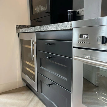 Moderne keuken met zwarte kasten met de Cordero Black Knob van yessential.nl in matzwart, ingebouwde wijnkoeler, roestvrijstalen oven met 8:34, gespikkeld granieten aanrecht, glaswerk in de bovenkast en koffiezetapparaat op het aanrecht.
