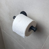 Een rol toiletpapier hangt aan de Tino Black toiletrolhouder van yessential.nl, gemonteerd tegen een lichtgrijs betegelde badkamermuur voor een moderne touch.
