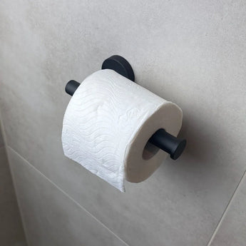 Een rol toiletpapier hangt aan de Tino Black toiletrolhouder van yessential.nl, gemonteerd tegen een lichtgrijs betegelde badkamermuur voor een moderne touch.