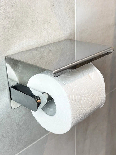 Een rol toiletpapier is gemonteerd op een Tazio Chrome (No Drill) houder van yessential.nl, met een plat, rechthoekig deksel in een betegelde badkamer.