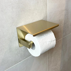 Een rol toiletpapier zit op de Tazio Gold wc-rolhouder van yessential.nl, elegant gemonteerd op een lichtgrijs betegelde badkamermuur.
