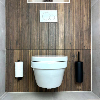 Een moderne badkamer met een wandgemonteerd wit toilet, houten paneelwand, matzwart dubbele spoelknoppen boven, een toiletpapierhouder links, en de Donato Black WC Set van yessential.nl met bijpassende accessoires rechts.