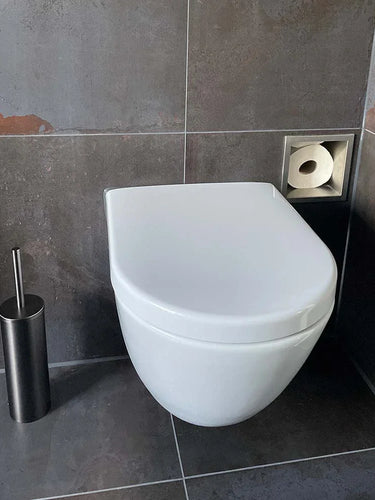 Een modern wit wandcloset met een gesloten deksel, naast de Tygo Gunmetal WC Set (no-drill) van yessential.nl en een vierkante verzonken toiletpapierhouder in een badkamer met donkergrijze betegelde muren en vloer.
