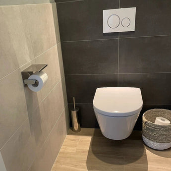 Een moderne badkamer met de Tazio Chrome toiletpapierhouder van yessential.nl, een wandcloset met dual-flush knoppen, een toiletborstel en een geweven mand op een lichte houten vloer en betegelde muren.