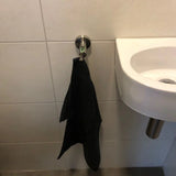 Een zwarte handdoek hangt aan een Justo RVS haak van yessential.nl aan een betegelde badkamermuur naast een kleine witte wastafel met een metalen afvoerpijp.