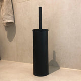 De Delo Black WC Set (no-drill) van yessential.nl, met een zwarte toiletborstel met cilindrische houder, staat op een beige badkamervloer tegen betegelde muren-een stijlvol matzwart accessoire voor eenvoudige installatie zonder boren.