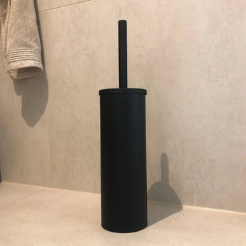 De Delo Black WC Set (no-drill) van yessential.nl, met een zwarte toiletborstel met cilindrische houder, staat op een beige badkamervloer tegen betegelde muren-een stijlvol matzwart accessoire voor eenvoudige installatie zonder boren.