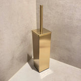 De Velasco Gold toiletborstel van yessential.nl, vrijstaand met houder, staat in de hoek van een badkamer met beige betegelde muren en vloer.