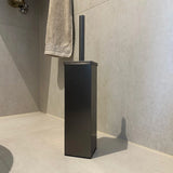 De Gio Gunmetal WC Set (zonder boren) van yessential.nl - een moderne, vierkante zwarte toiletborstelhouder met bijpassende handgreep - rust op lichte tegels in de buurt van een handdoek, ideaal voor een stijlvolle toiletaccessoireset zonder boren.
