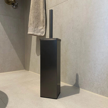 De Gio Gunmetal WC Set (zonder boren) van yessential.nl - een moderne, vierkante zwarte toiletborstelhouder met bijpassende handgreep - rust op lichte tegels in de buurt van een handdoek, ideaal voor een stijlvolle toiletaccessoireset zonder boren.
